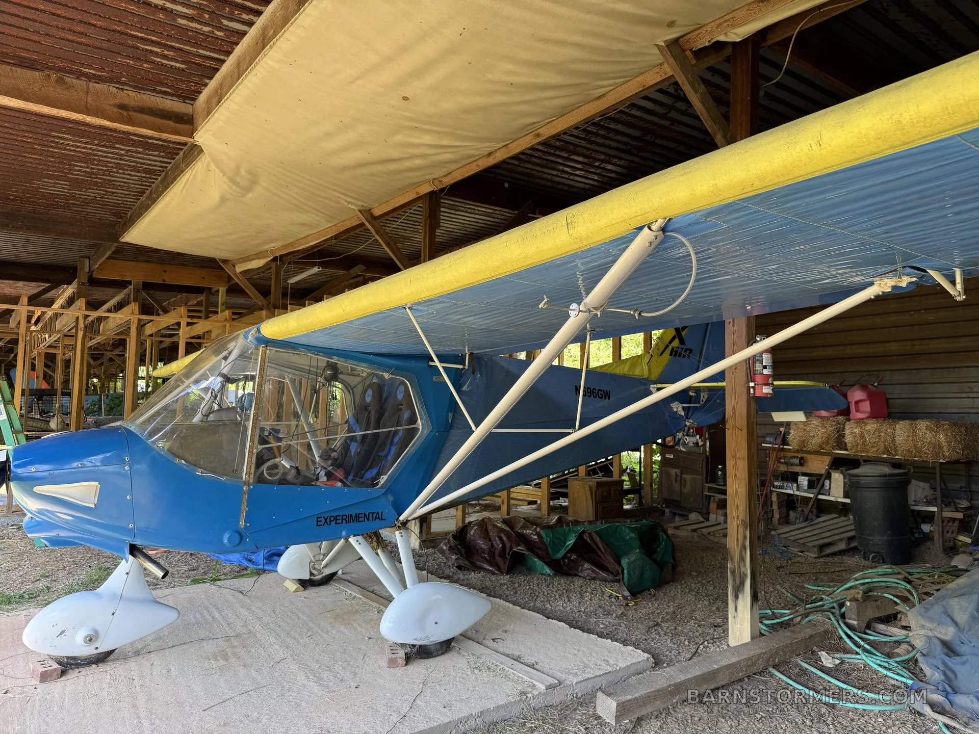 A blue and yellow experimental aircraft with white floats is parked inside a rustic shed.