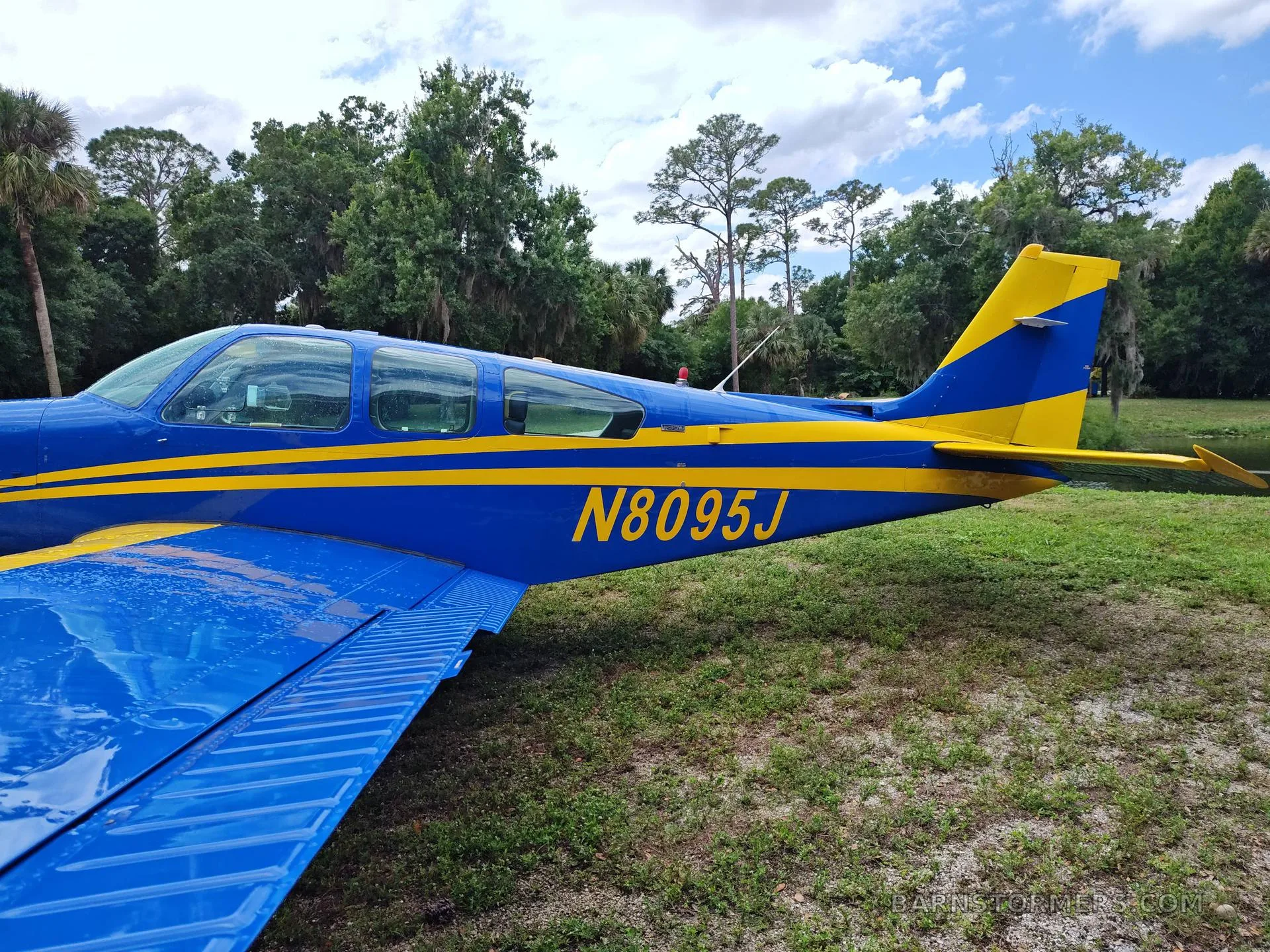 A close-up side view of the blue and yellow fuselage and tail of a single-engine aircraft, showing the registration N8095J.