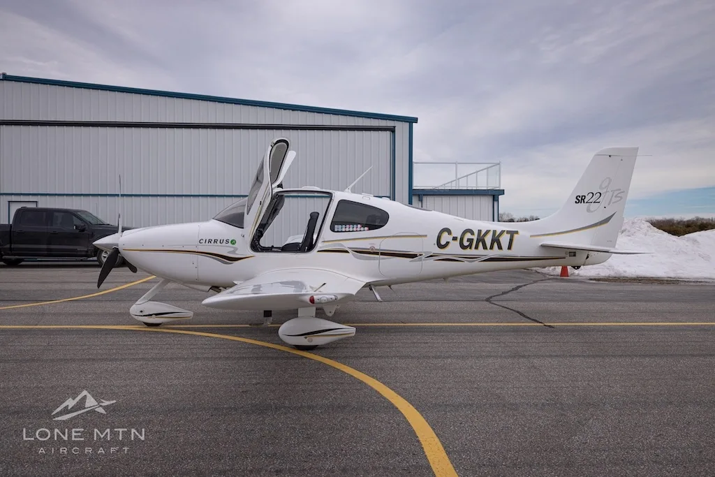 A side view of a white Cirrus SR22 aircraft with registration C-GKKT, doors open, parked on an airfield.