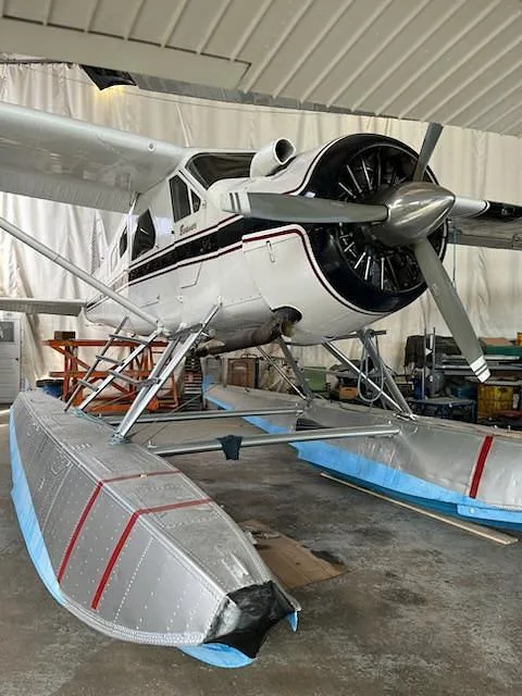 A white De Havilland Beaver seaplane with blue and silver floats is parked inside a hangar, viewed from the front-right.