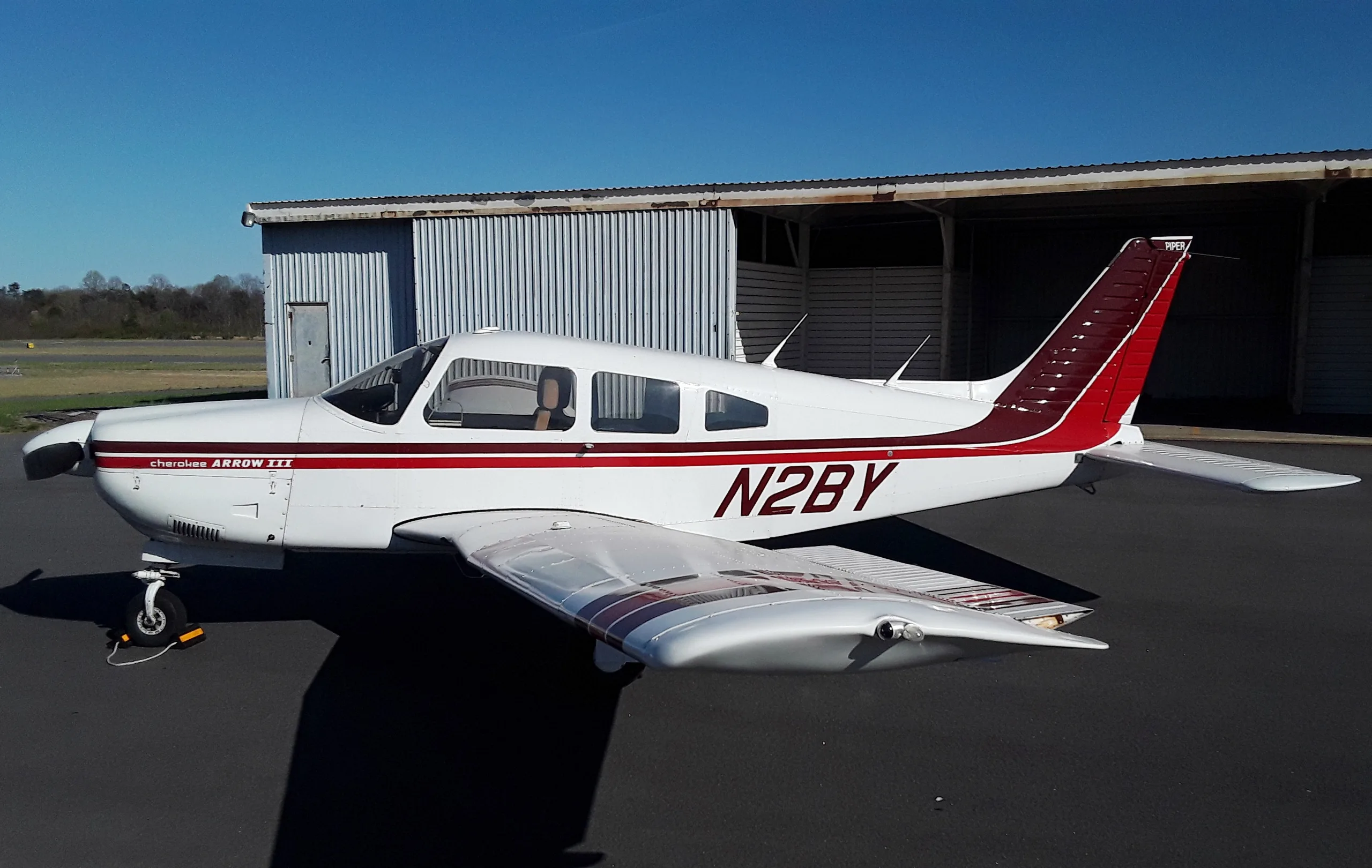 A white Piper Cherokee Arrow III with red stripes and tail, registration N2BY, parked on an asphalt ramp next to a hangar.