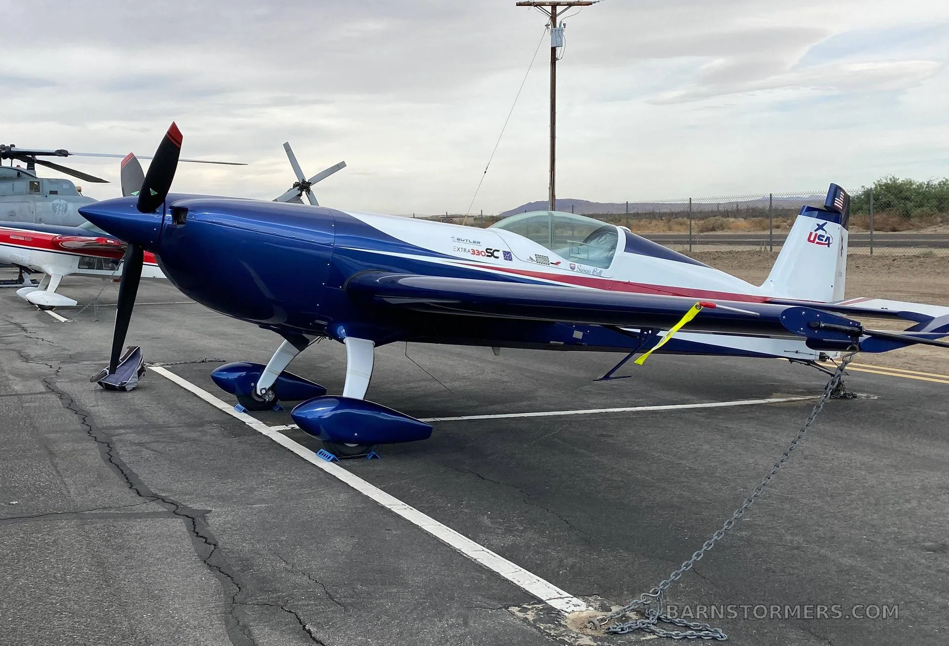 An Extra 330SC aircraft with blue, white, and red stripes is parked and chained on an airfield tarmac.