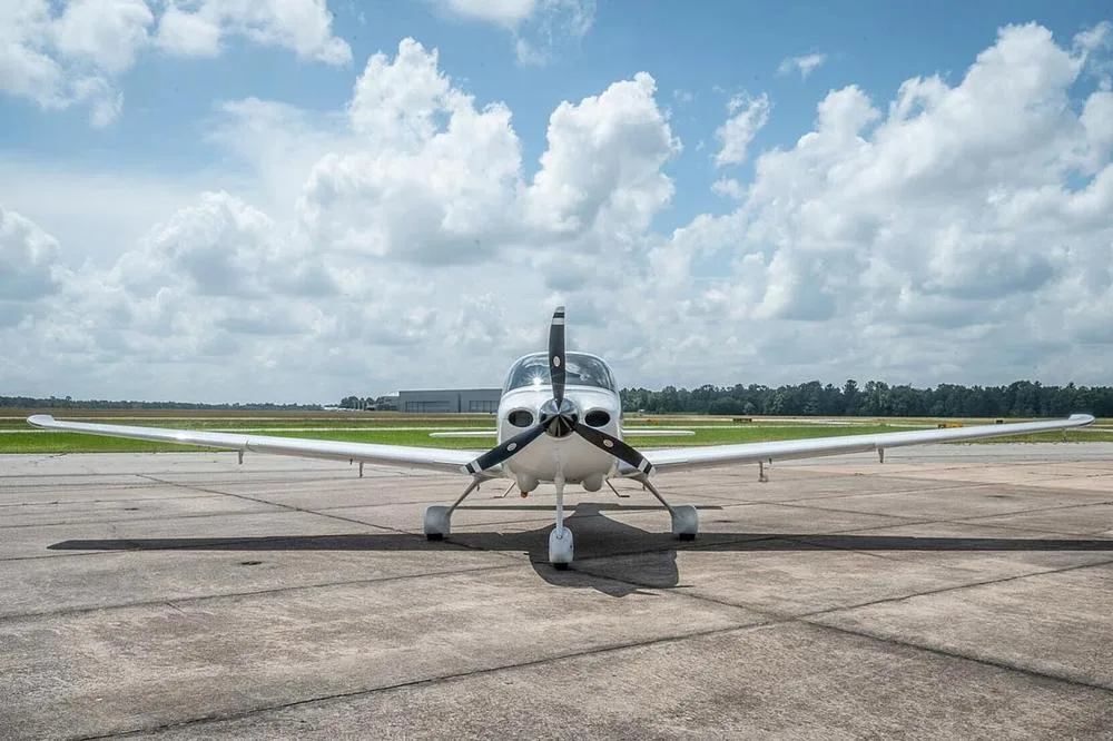 A head-on view of a white Cirrus SR22 aircraft with a three-bladed propeller, parked on an airfield.