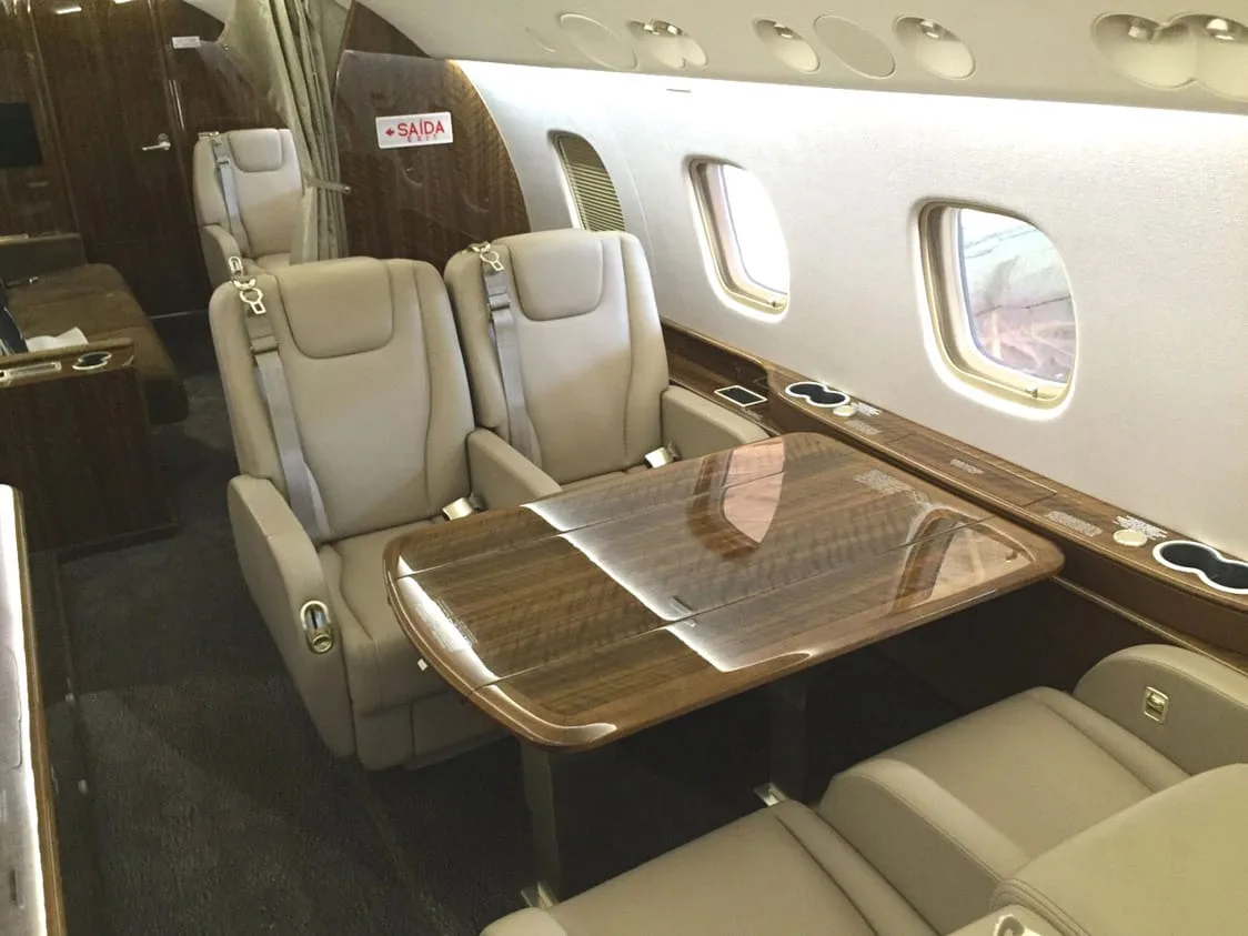The luxurious interior of a private jet cabin, with beige leather seats and a polished wooden table by the windows.