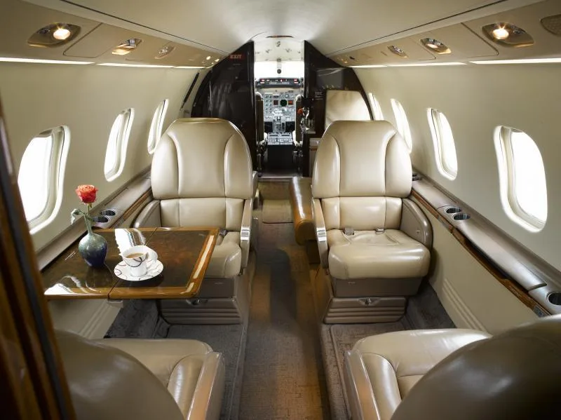 View down the aircraft cabin aisle showing tan leather seats, a polished wood table with a rose and coffee cup, and the cockpit visible in the distance.