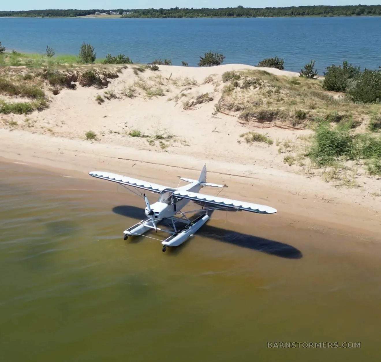 A white seaplane with floats is partially in the water, parked on a sandy beach next to a large lake.