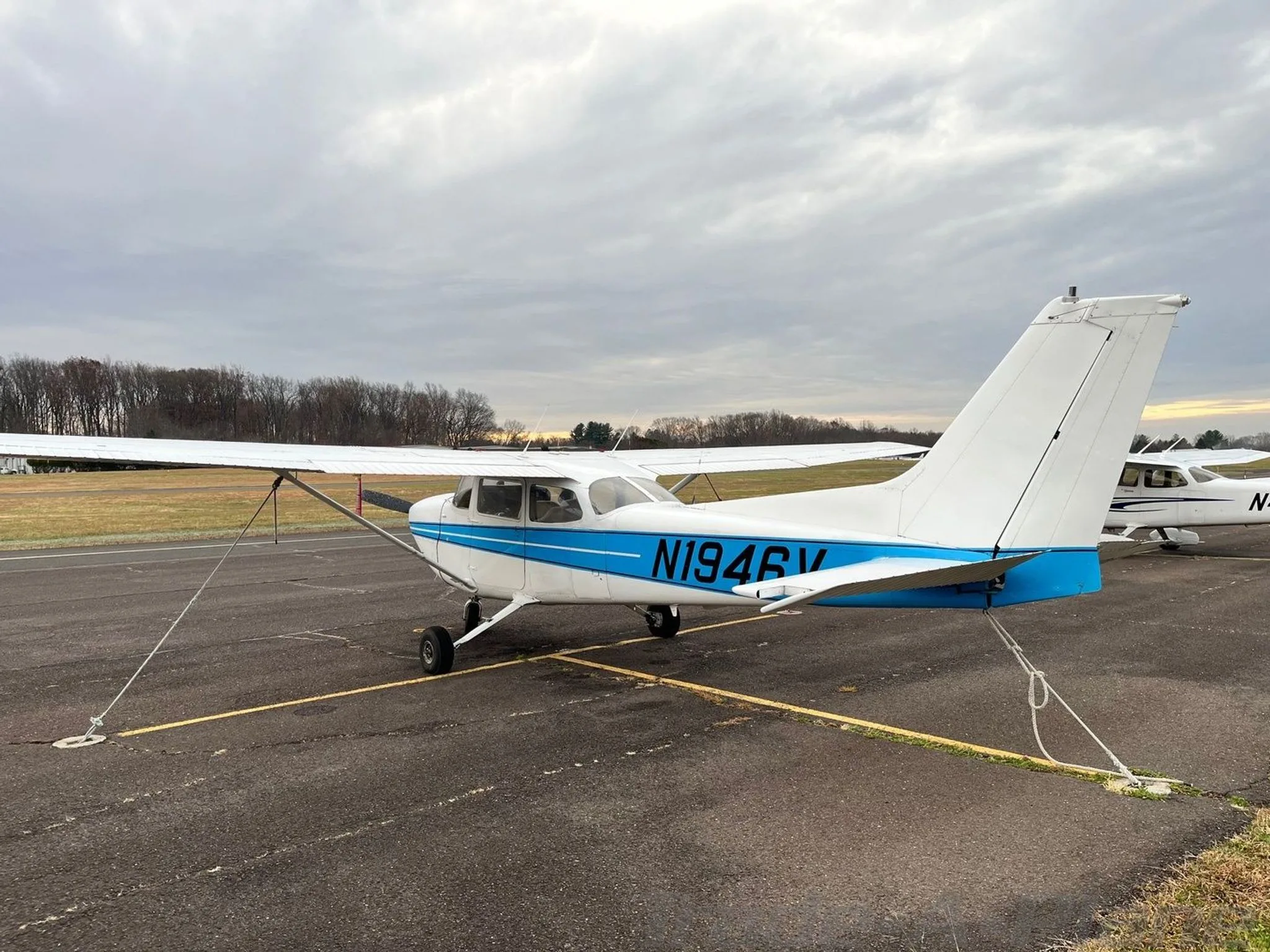 A side view of the N1946V aircraft, a white plane with blue stripes, tied down on an airfield.