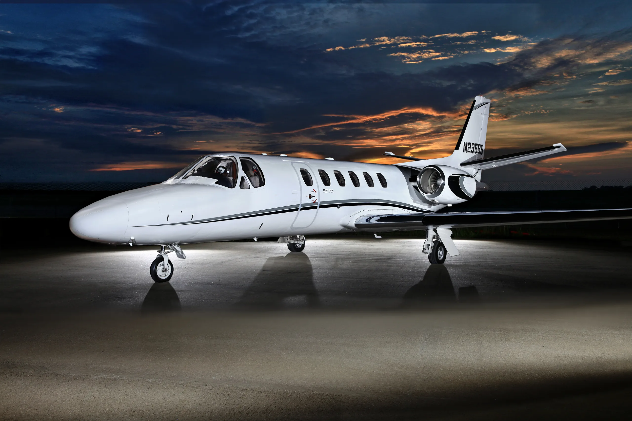 A white business jet with black and grey stripes, registration N235BS, parked on an tarmac at dusk with a dramatic sky.