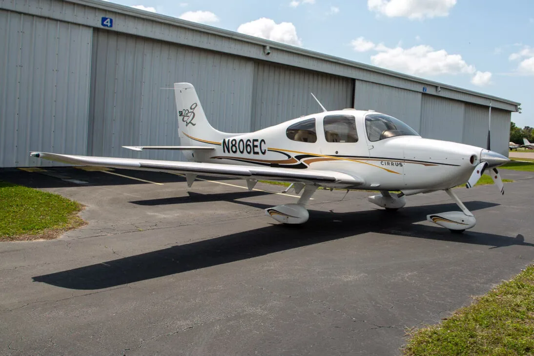 A white Cirrus SR22 aircraft with black and gold stripes, registration N806EC, parked on an asphalt tarmac in front of a hangar.