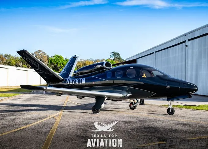 A dark blue Cirrus Vision Jet (N876TM) is parked on an asphalt ramp next to a white hangar under a clear blue sky.