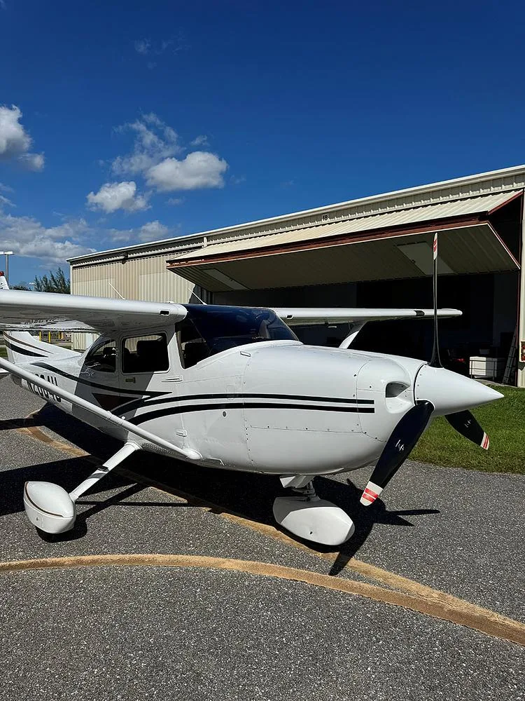Front-right view of a white Cessna 182 Skylane with black and brown stripes, parked on an asphalt ramp in front of an open hangar.