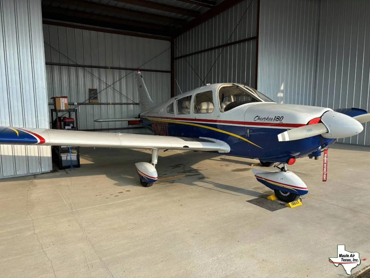A white and blue Piper Cherokee 180 aircraft with red and yellow stripes is parked inside a hangar.
