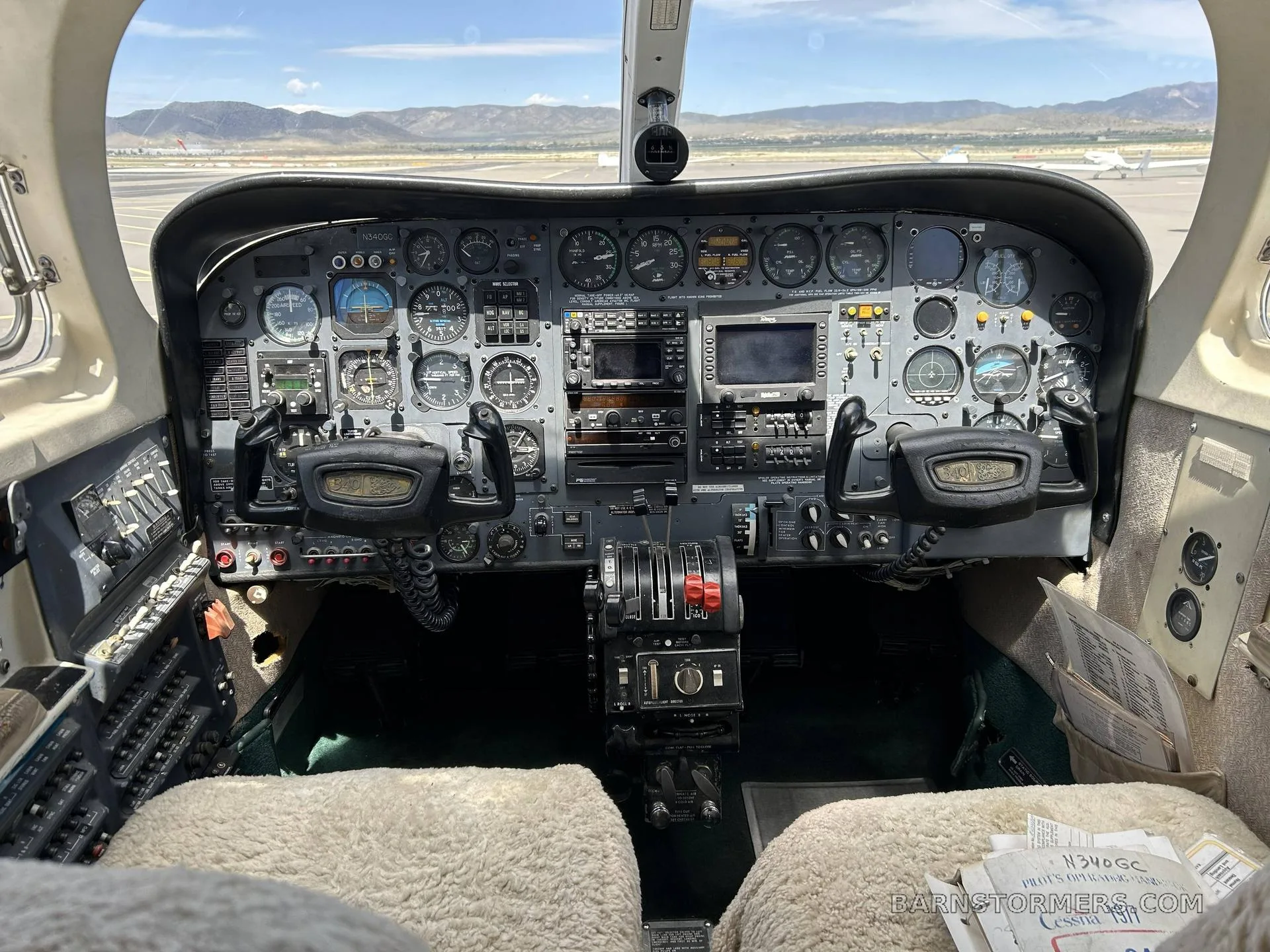 The cockpit of a twin-engine aircraft, showing the instrument panel, yokes, and center console, with an airfield visible through the windshield.