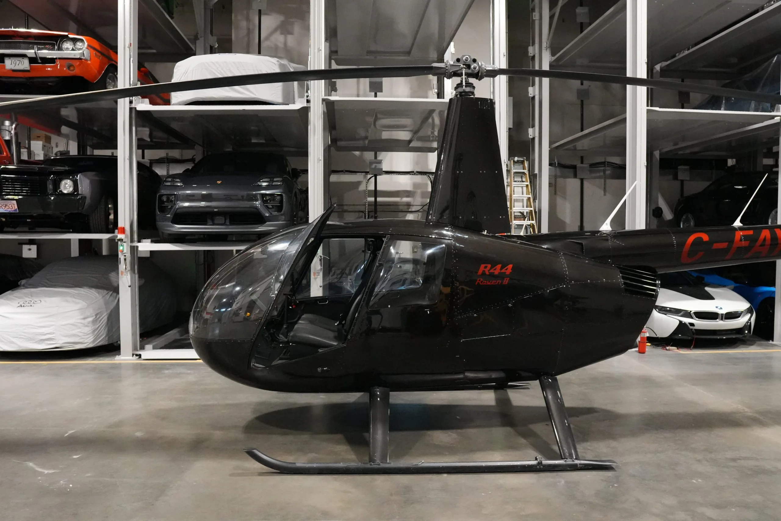 A black Robinson R44 Raven II helicopter (C-FAYW) with an open door, parked in a hangar surrounded by stored cars.