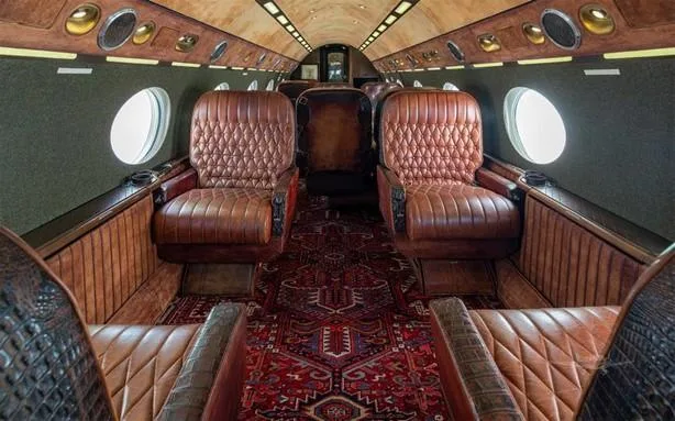 The luxurious aircraft cabin features brown quilted leather seats, a patterned red rug, and wood paneling.