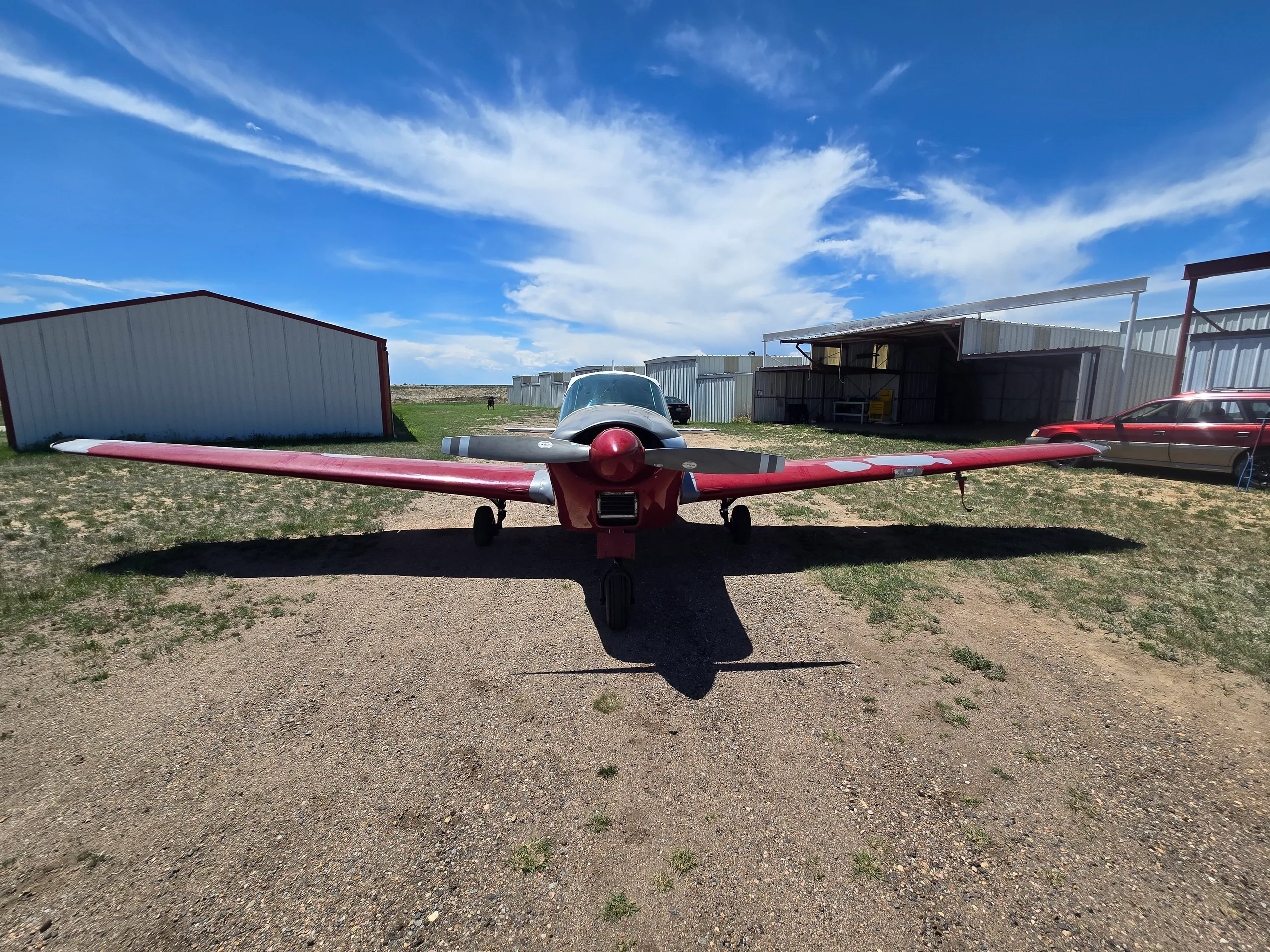 A red and silver single-engine aircraft with a propeller, viewed from the front, parked on a dirt path between hangars.