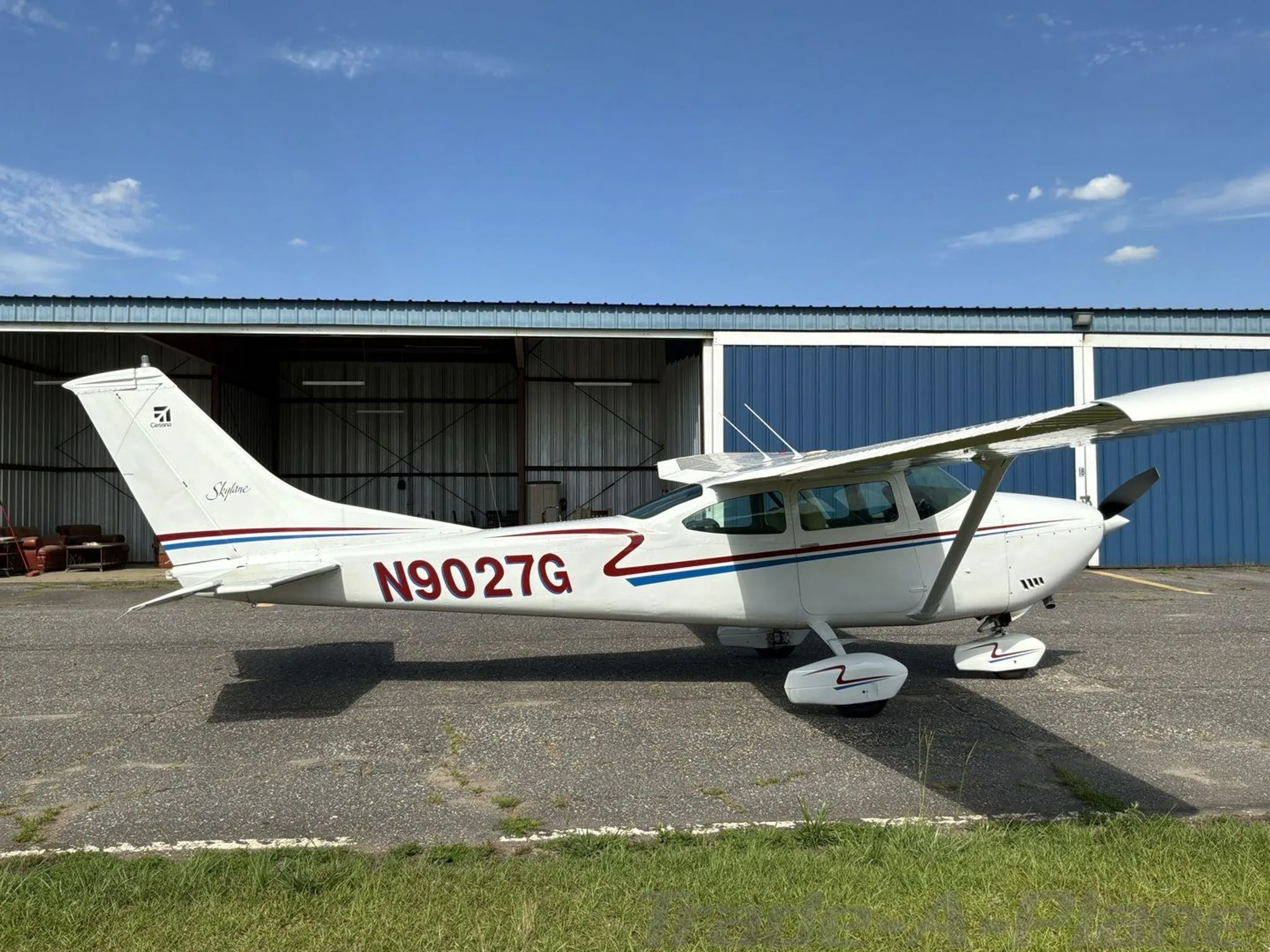 A white Cessna 182N Skylane with red and blue stripes, registration N9027G, parked on an asphalt ramp next to a blue hangar.