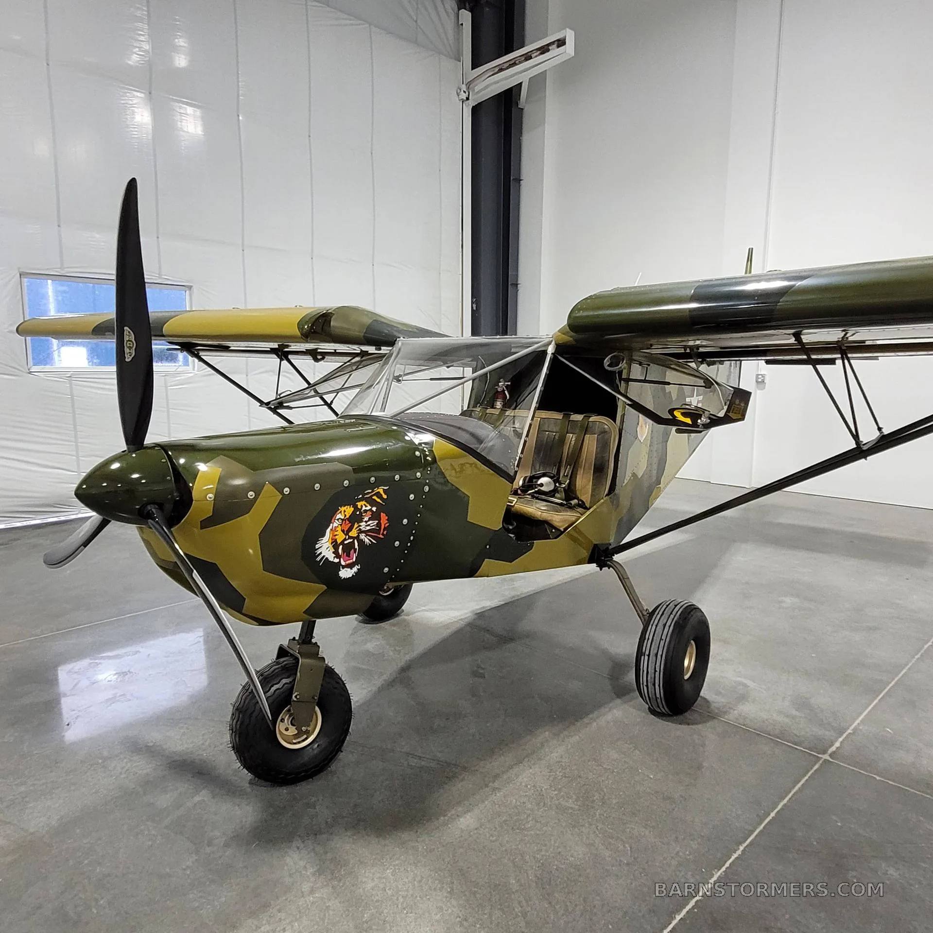 A small, single-engine, high-wing aircraft with a green and brown camouflage paint scheme and a tiger head decal on the nose, parked in a hangar.