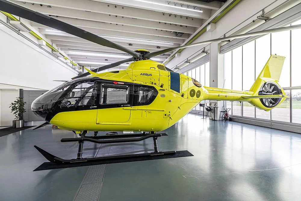 A bright yellow Airbus H135 helicopter with black accents sits on a hangar floor, viewed from the front-right.
