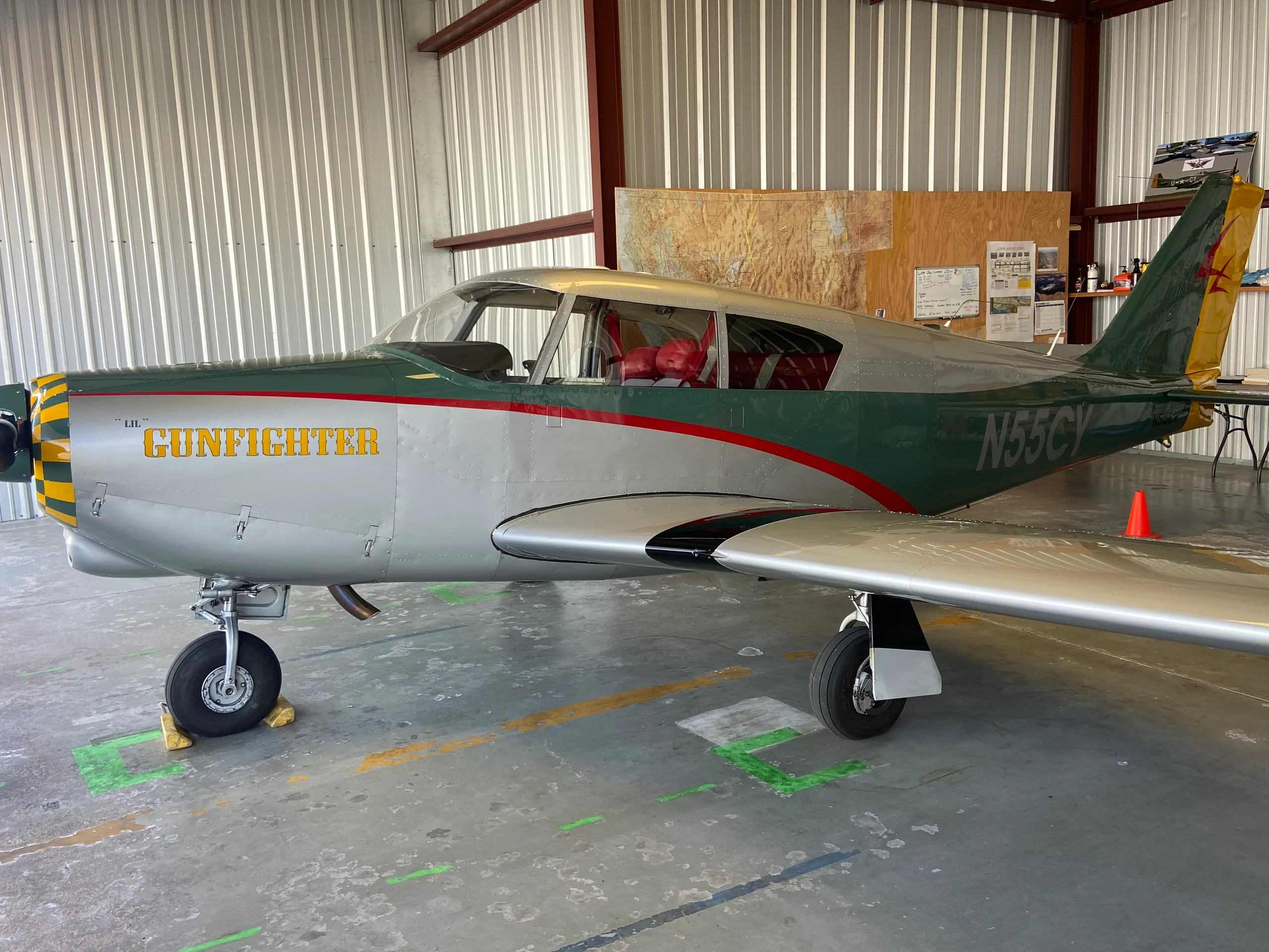 A silver and green single-engine propeller aircraft named "GUNFIGHTER" is parked inside a hangar.