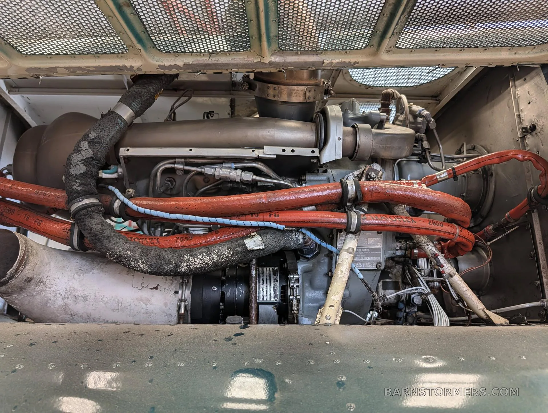 A detailed view of a helicopter's engine compartment, showing the engine, exhaust, and various red-sleeved hoses.