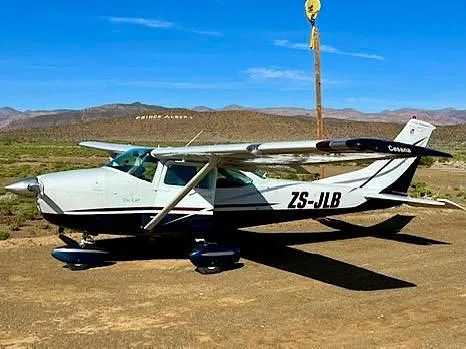 A white and blue Cessna ZS-JLB aircraft is parked on a dirt airstrip with mountains in the background under a clear blue sky.
