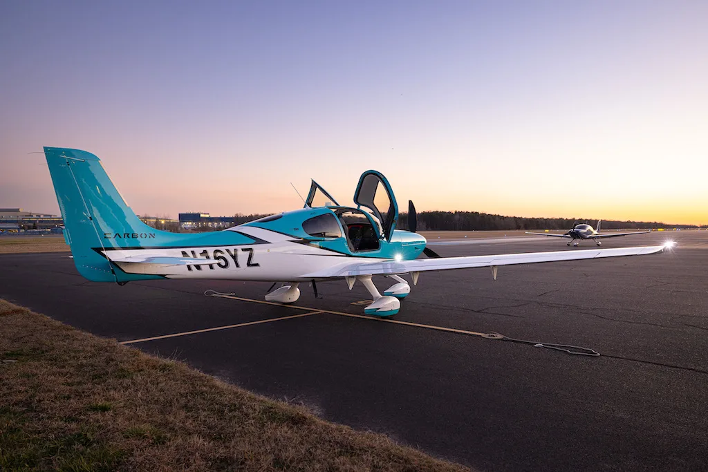 A turquoise and white Cirrus SR22 aircraft with its doors open, parked on an airfield with another plane in the distance at dusk.