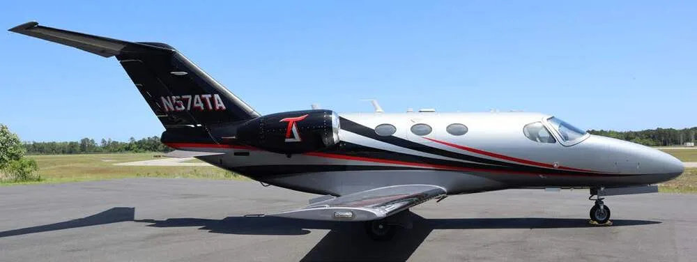 A silver, black, and red Cessna Citation Mustang jet, tail number N574TA, parked on an tarmac with a grassy field in the background.