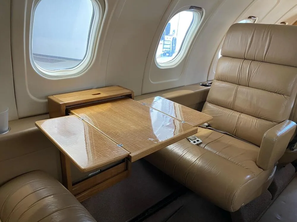 Interior view of an aircraft cabin showing a tan leather seat and a wooden fold-out table extended.