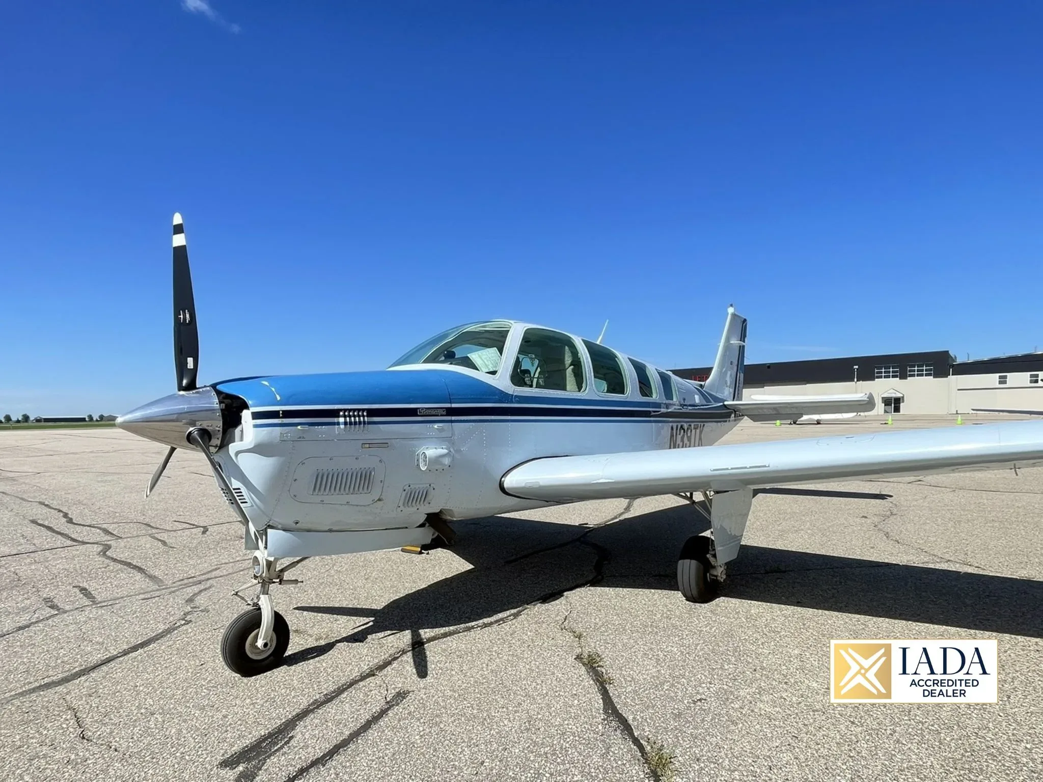 A blue and white single-engine propeller aircraft, registration N39TK, is parked on an asphalt tarmac under a clear blue sky.