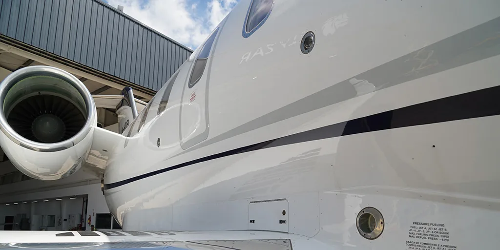A close-up view of the white fuselage and engine of a business jet, showing fuel port details and a dark blue stripe, parked near a hangar.