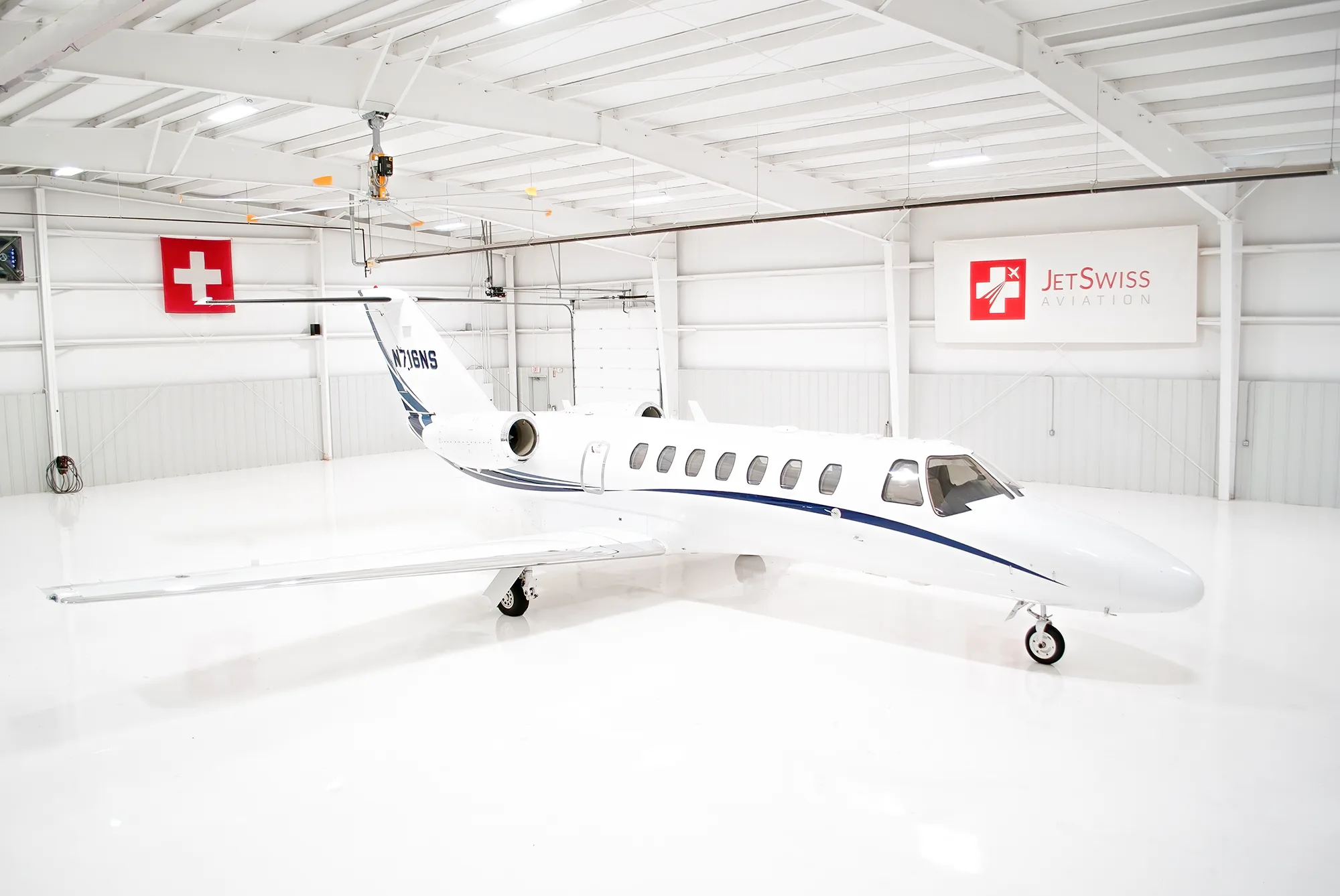 A white Cessna Citation business jet with blue stripes, registration N716NS, parked inside a brightly lit hangar.