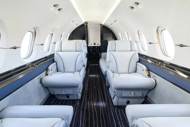 The luxurious interior cabin of a business jet with light grey leather seats and dark patterned carpet.