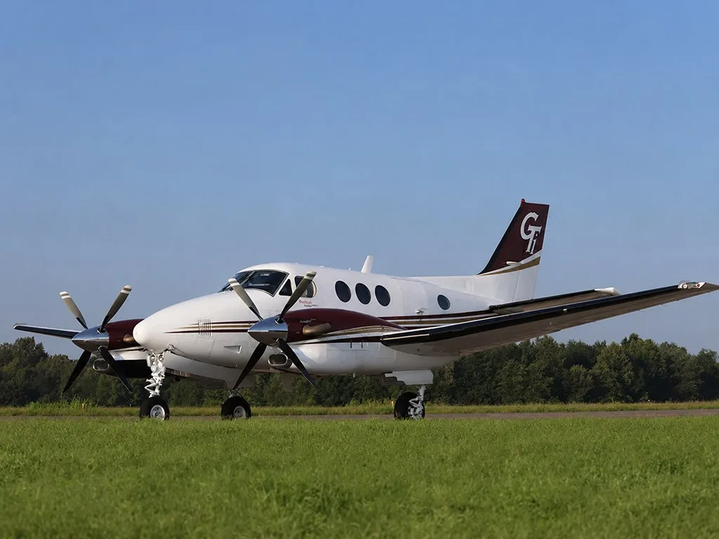 A white and maroon Beechcraft King Air 350 with a 'GT' logo on the tail, parked on a grassy airfield under a clear blue sky.