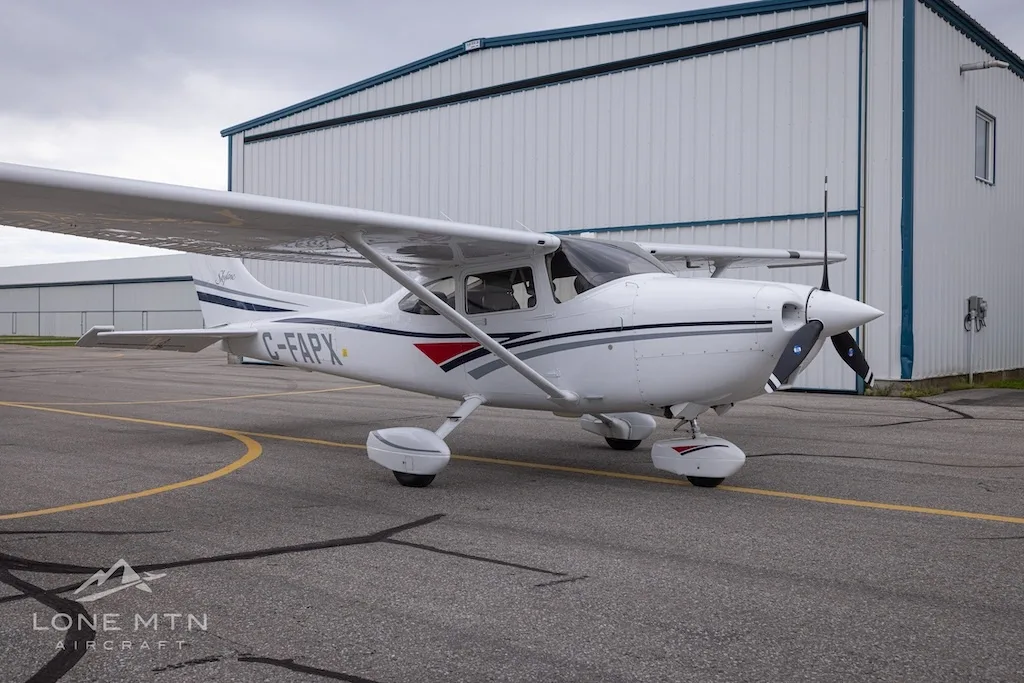 A white Cessna 182 Skylane with grey and blue stripes, registration C-FAPX, parked on an asphalt ramp with a hangar in the background.