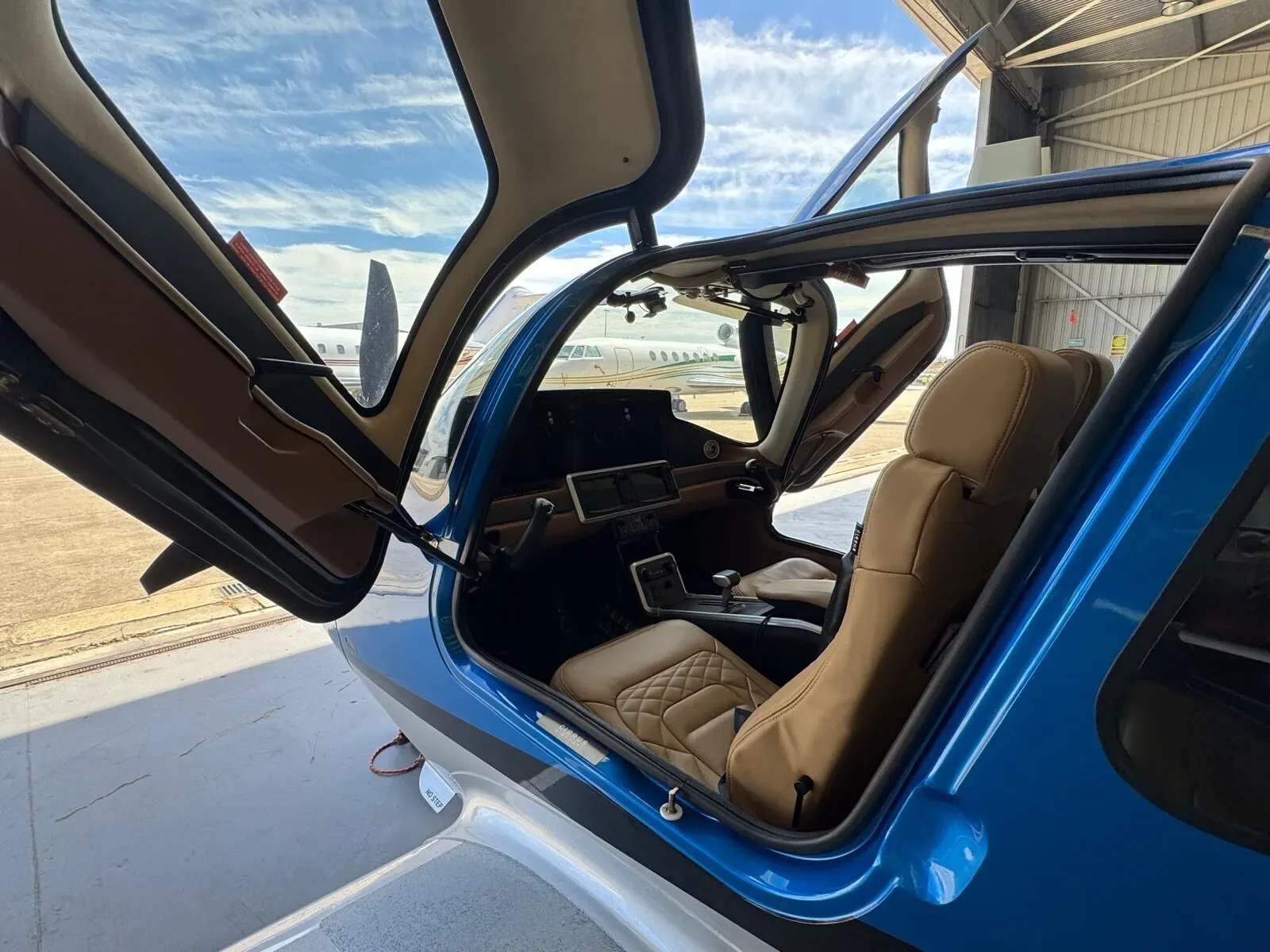 The tan leather interior and cockpit of a blue Cirrus SR22 aircraft with its doors open, viewed from the right side.
