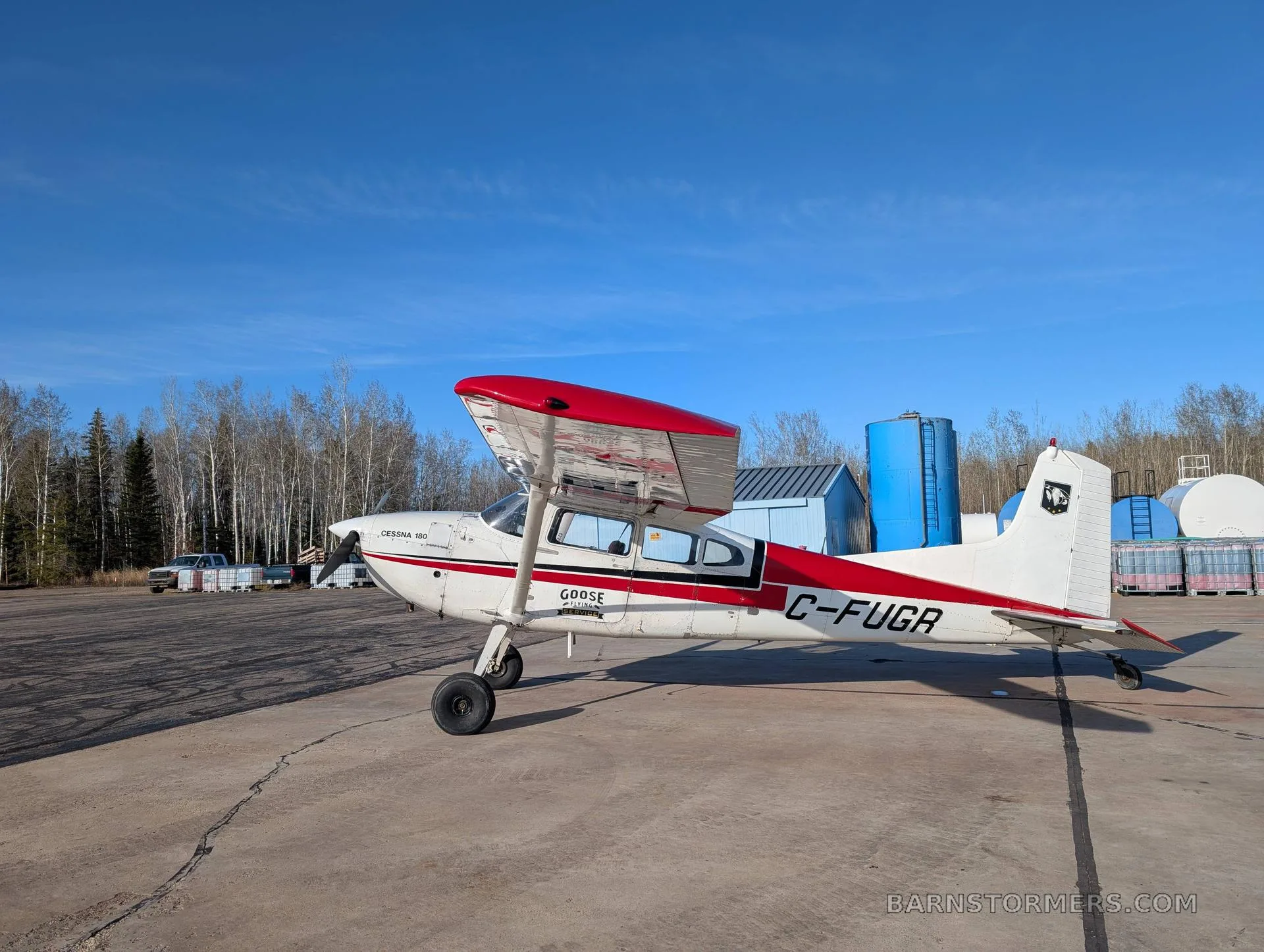 A white and red Cessna 180 (C-FUGR) is parked on a concrete tarmac with blue storage tanks and trees in the background.
