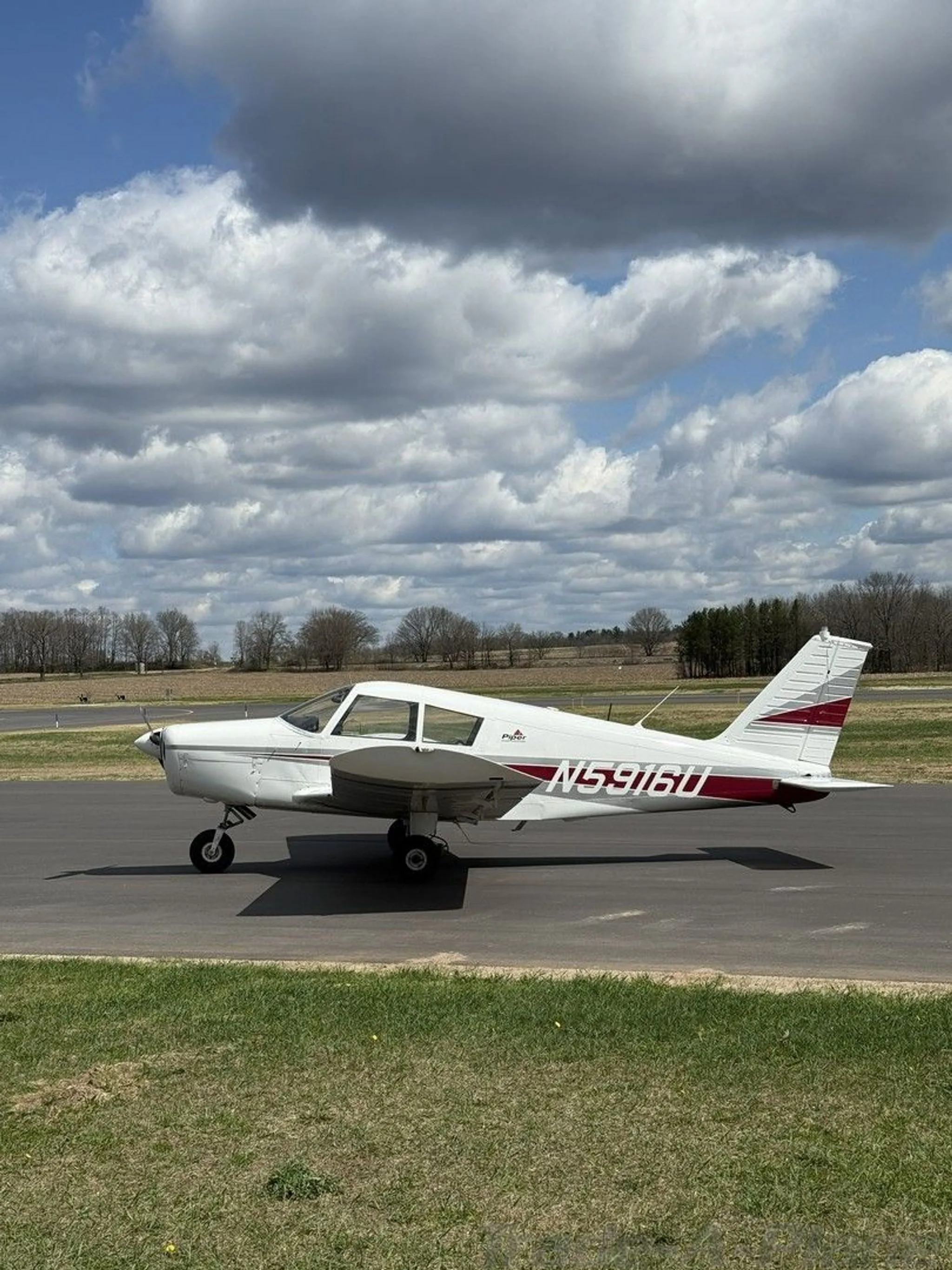 A white Piper Cherokee aircraft with red and grey stripes on an airfield under a partly cloudy sky.