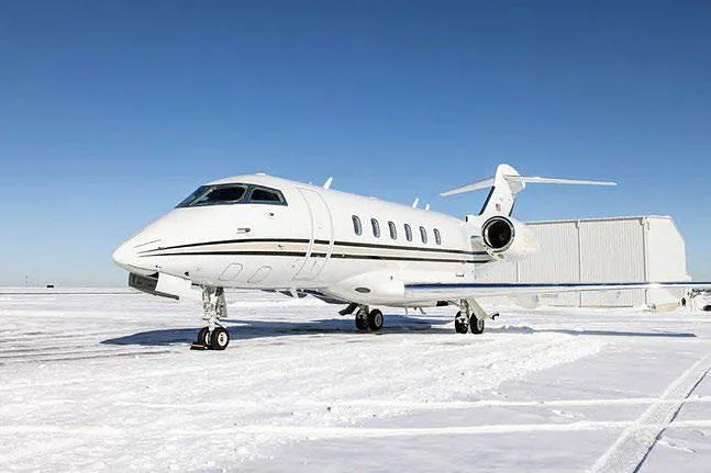 A white Bombardier Challenger business jet with black and gold stripes on a snowy tarmac under a clear blue sky.