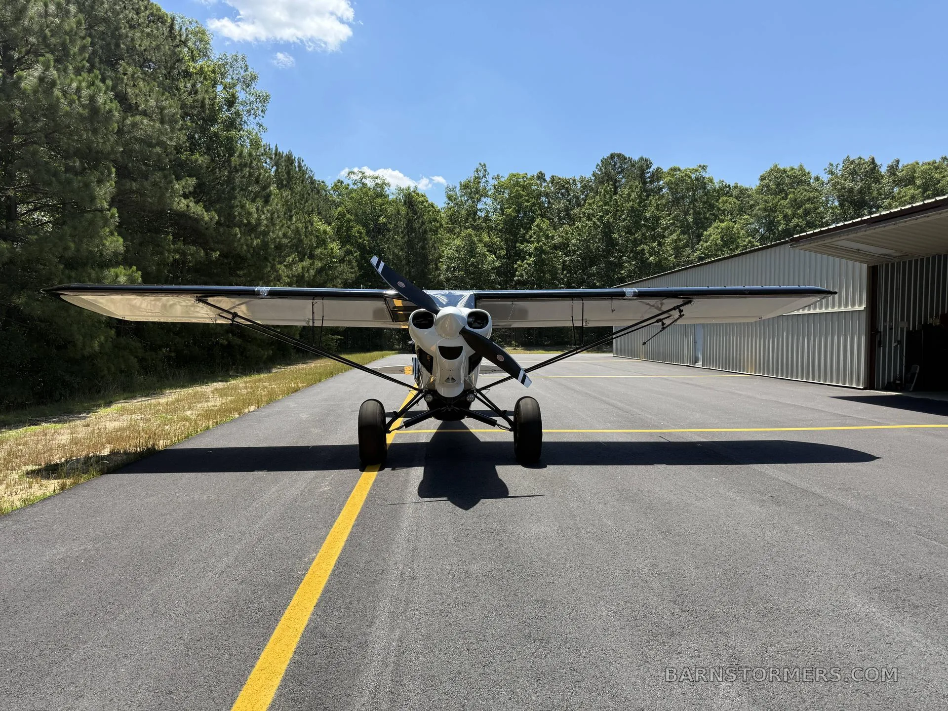 A head-on view of a white Carbon Cub FX-3 aircraft with a black propeller is parked on an asphalt runway in front of a hangar and trees.