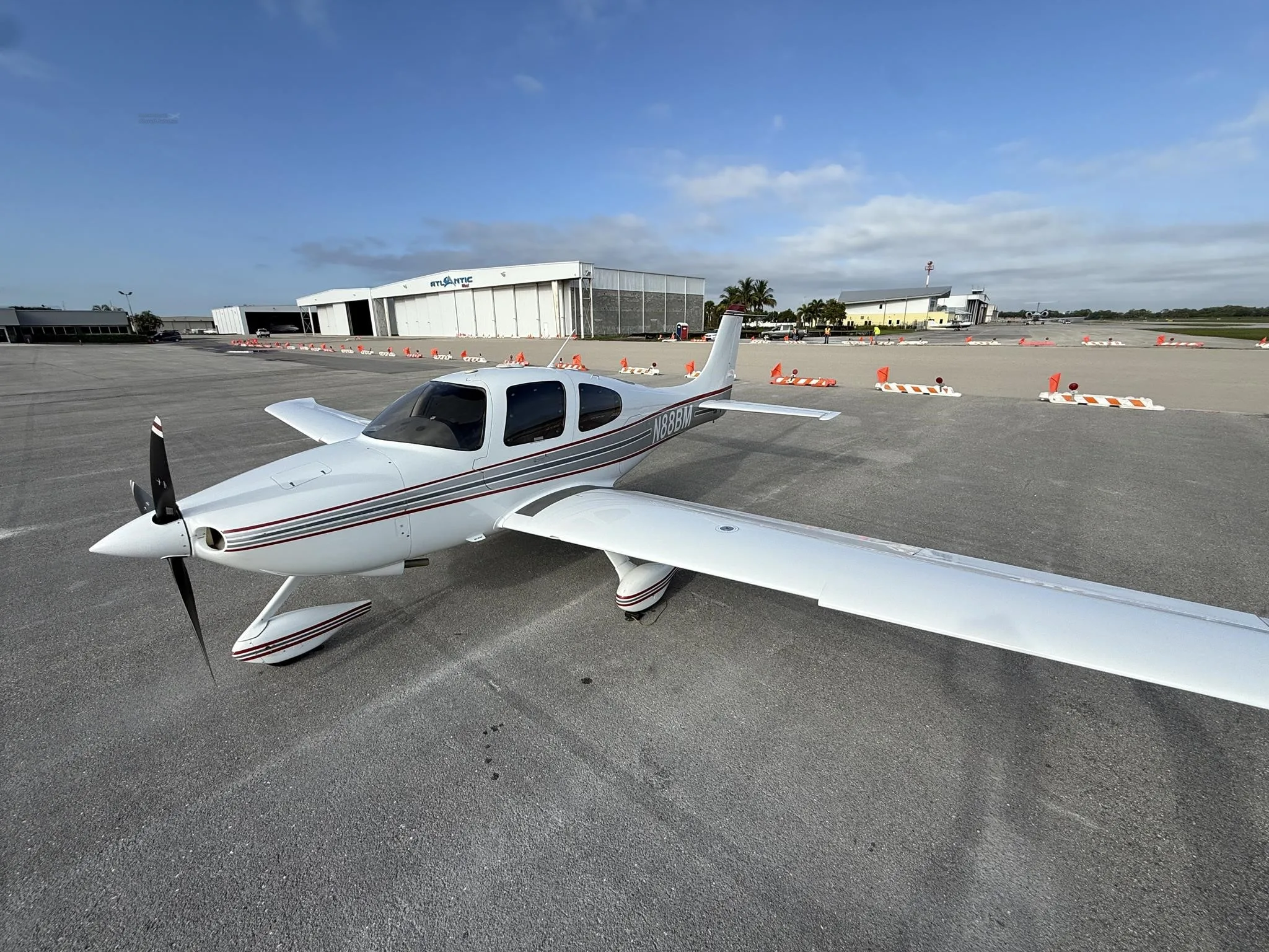 A white Cirrus SR22 aircraft with red and gray stripes is parked on an airport tarmac under a blue sky.