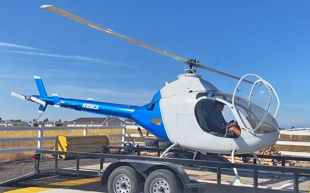 A blue and white helicopter (N129CA) with a pilot in the cockpit, loaded on a trailer under a clear sky.