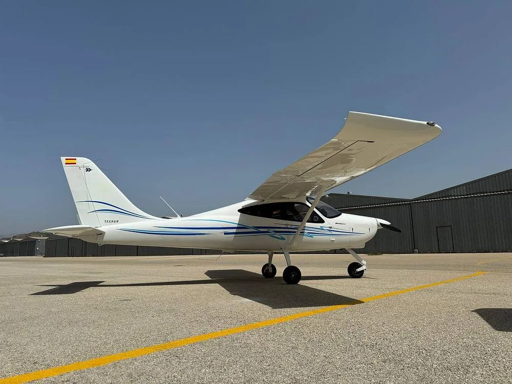 A white Tecnam P2008 aircraft with blue stripes on the tail and fuselage, parked on an asphalt apron with hangars in the background.
