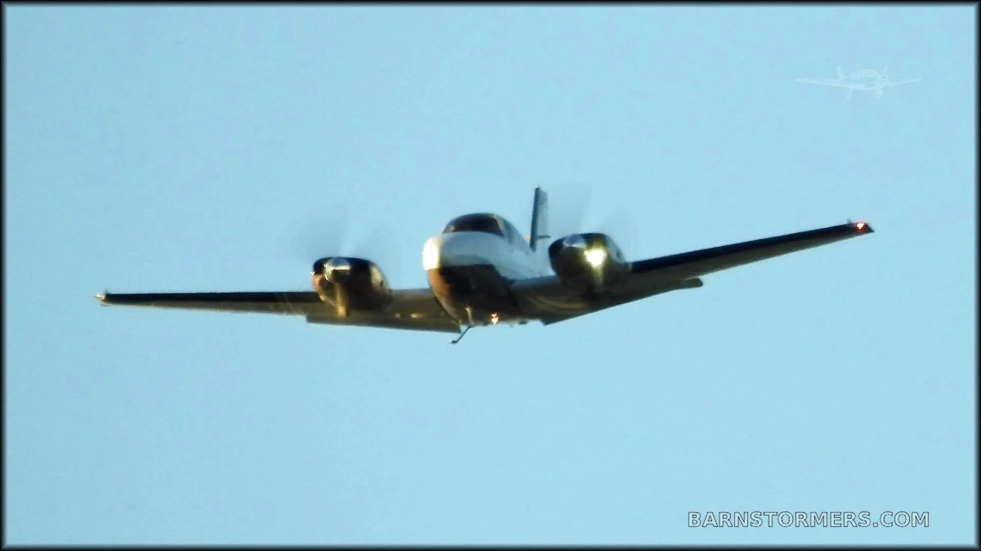 A twin-engine aircraft with a white nose and dark propellers is flying against a clear blue sky, viewed from the front.