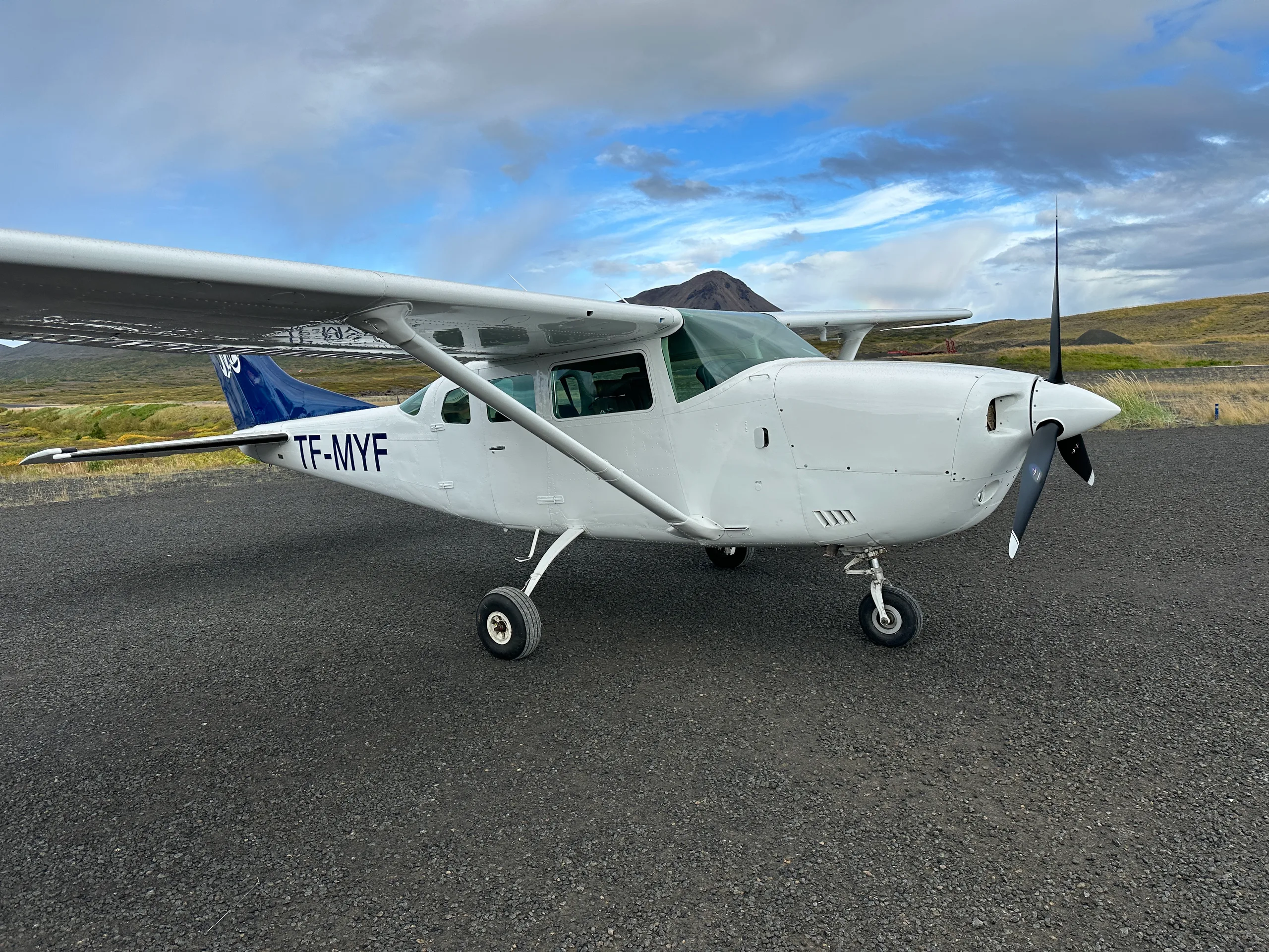 A white single-engine propeller aircraft with blue tail accents, registration TF-MYF, parked on a tarmac with a mountain in the background.