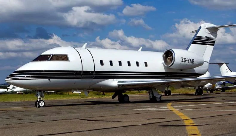 A white Bombardier Challenger business jet with black stripes and registration ZS-YAG on a tarmac under a cloudy sky.