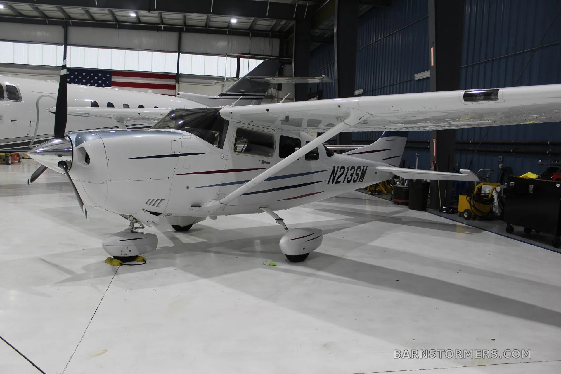 A white Cessna 210 Centurion (N213SM) with blue and red stripes, parked in a hangar, viewed from the front-left.