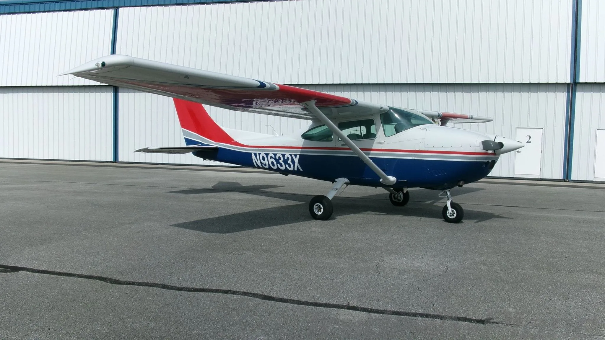 A white, blue, and red Cessna 182 Skylane with registration N9633X parked on an asphalt apron next to a white hangar.