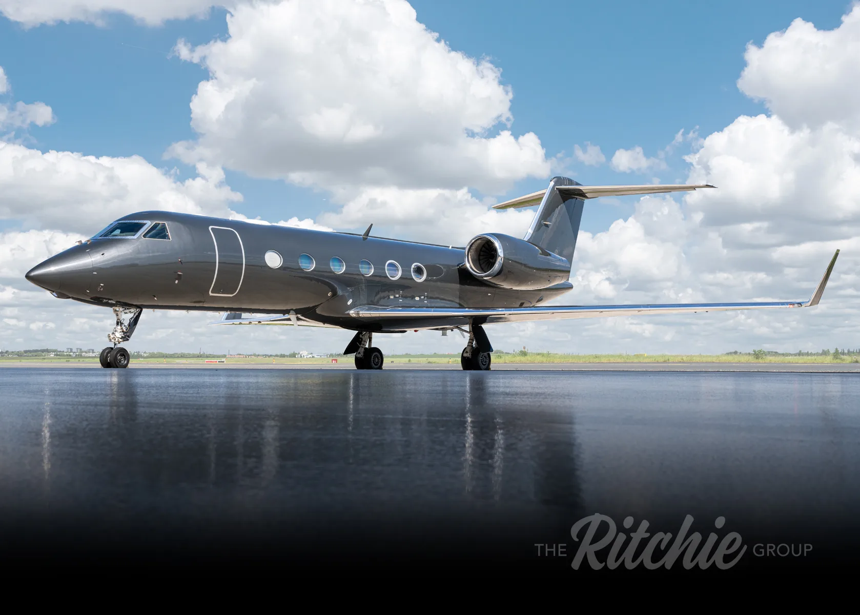 A dark grey Gulfstream G650 business jet parked on a tarmac under a partly cloudy sky, viewed from the front-left.