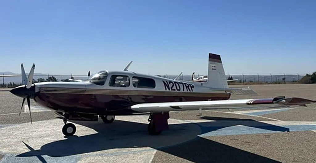 A white and maroon single-engine propeller aircraft, N207RP, is parked on an airfield with distant hills, viewed from the front-left.